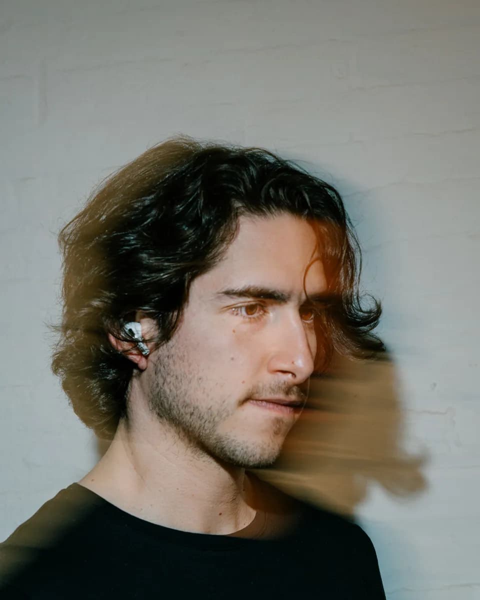 Portrait of a man with dark wavy hair and a wireless earbud against a white brick wall, hair blurred by motion