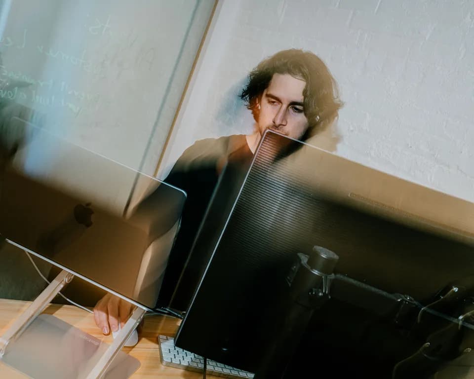 Man with dark wavy hair working at two monitors beside a white brick wall, shot with motion blur and reflections