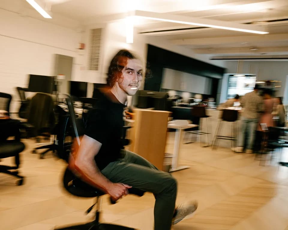 Man in a black tee and green trousers spinning on an office chair in a bright open office, blurred motion
