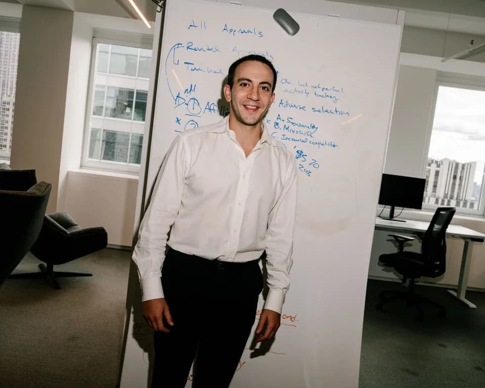 Smiling man in a white dress shirt standing in front of a whiteboard with blue handwriting, city windows behind him