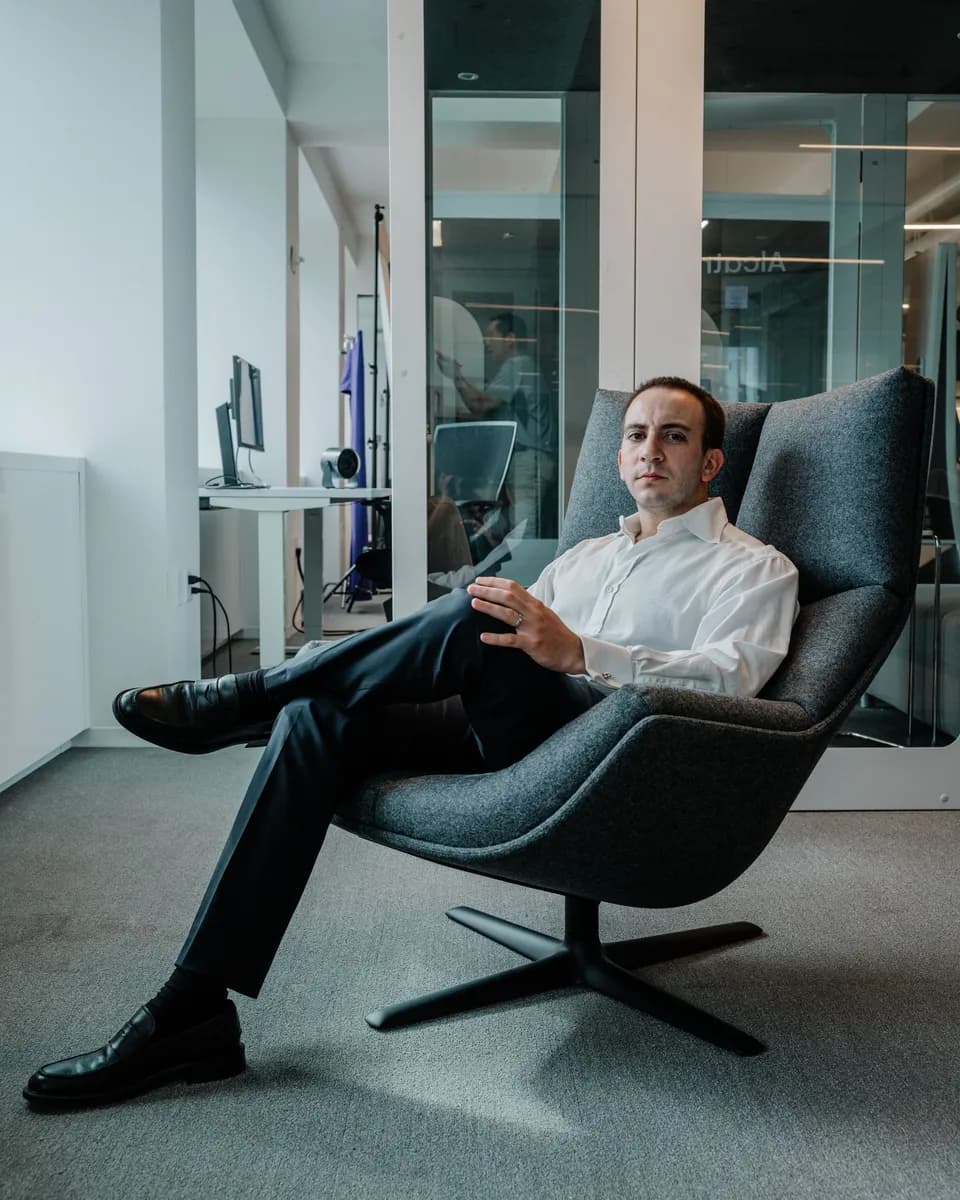 Man in a white dress shirt and dark trousers lounging in a grey swivel chair in a glass-walled office