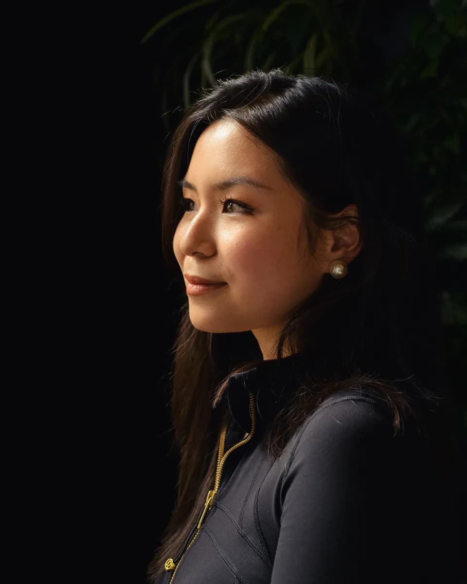 Side profile of a woman with long dark hair and pearl earrings, lit against a black background