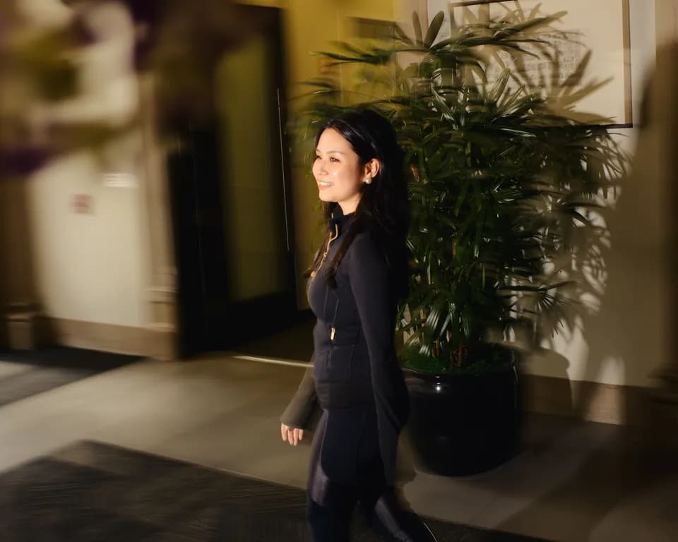 Portrait of a woman in a black zip jacket walking past a large potted palm in a warmly lit lobby