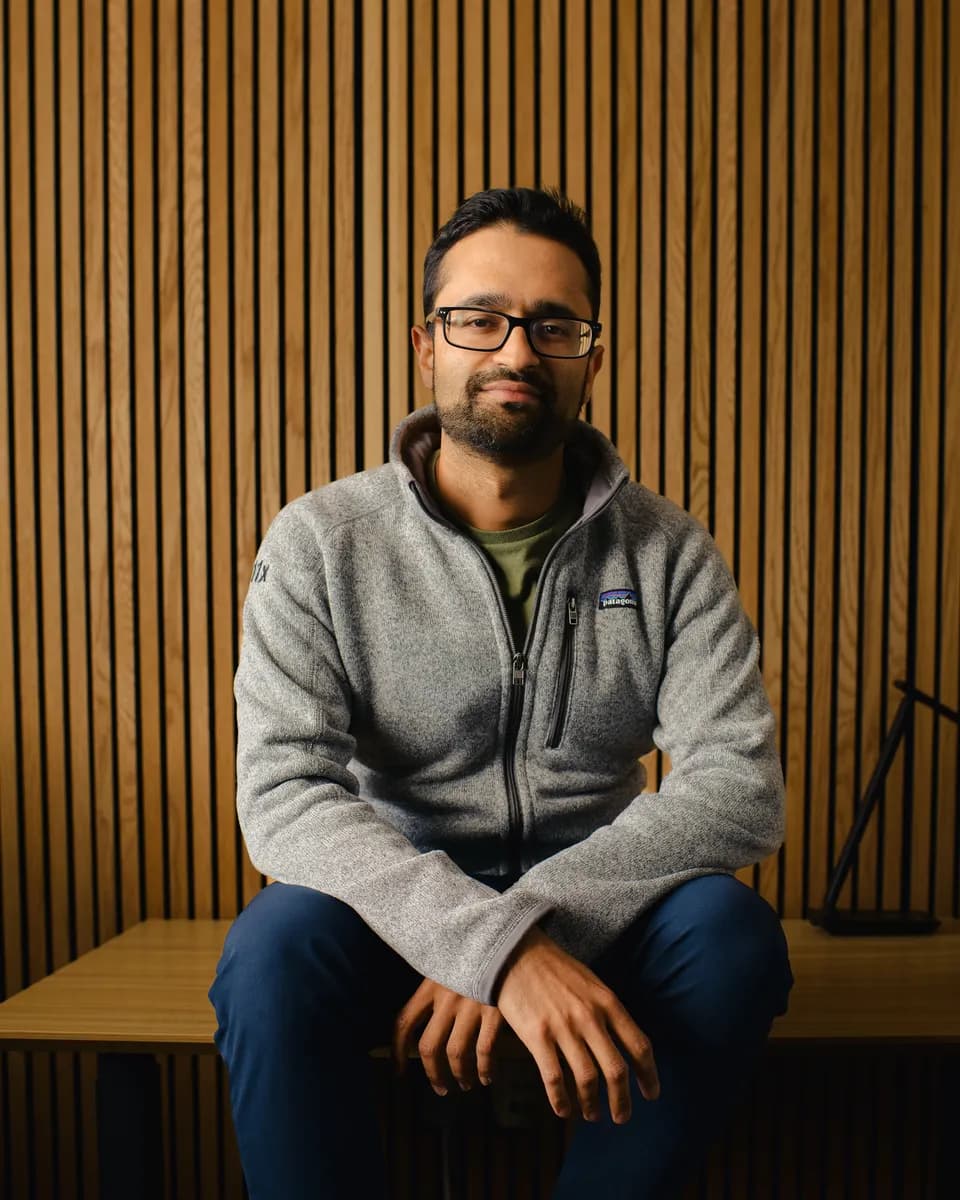 Brex Quitters Welcome portrait: a bearded man in glasses and a grey fleece sitting in front of a wood-slat wall