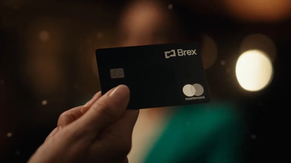 Hand holding up a black Brex Mastercard with a chip, with warm blurred lights behind