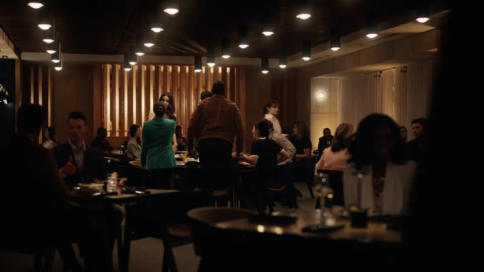 Wide, dim restaurant interior with pendant lights and diners at tables, a woman in a green jacket standing