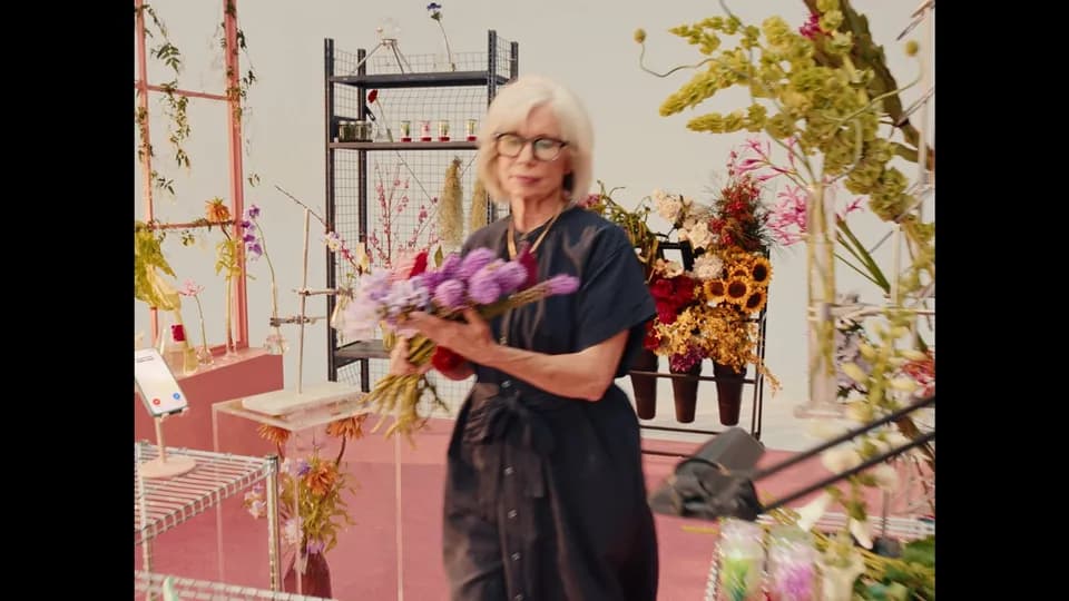 Older woman with glasses holding a bunch of purple flowers on a pink-floored florist set