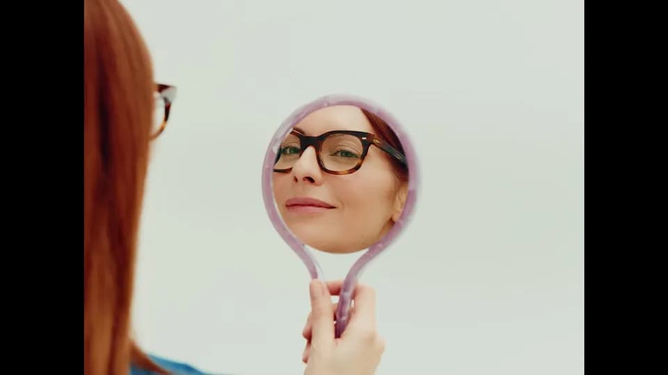 Woman in tortoiseshell glasses looking into a lilac hand mirror against a pale background