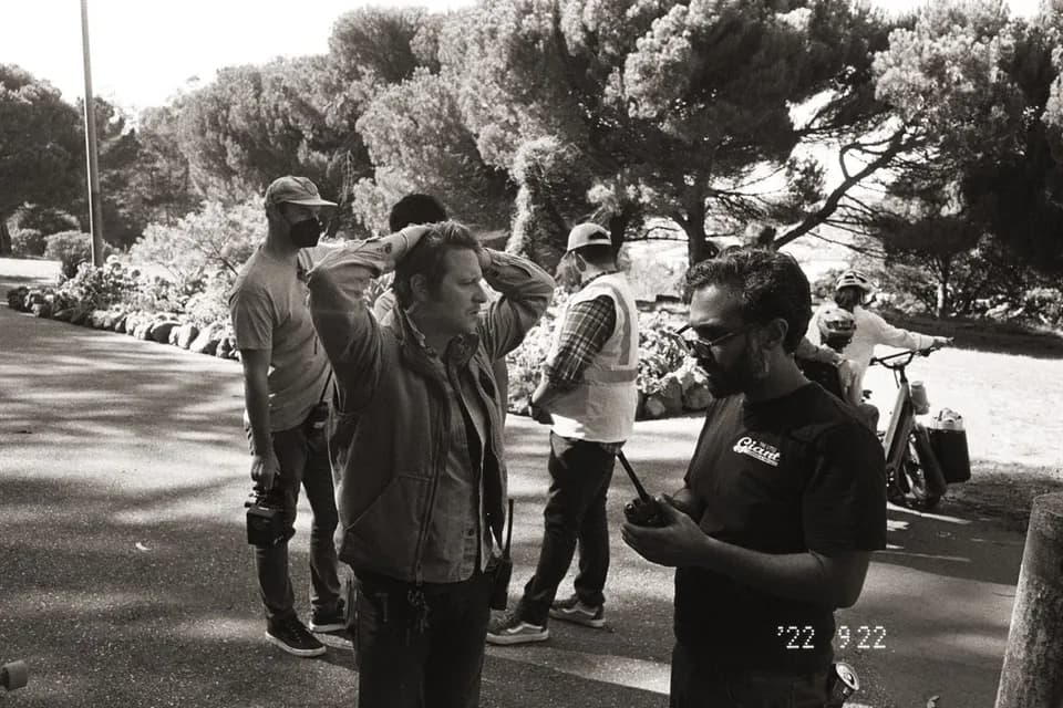 Black and white BTS photo of crew in a park, one man with hands on his head, another holding a radio