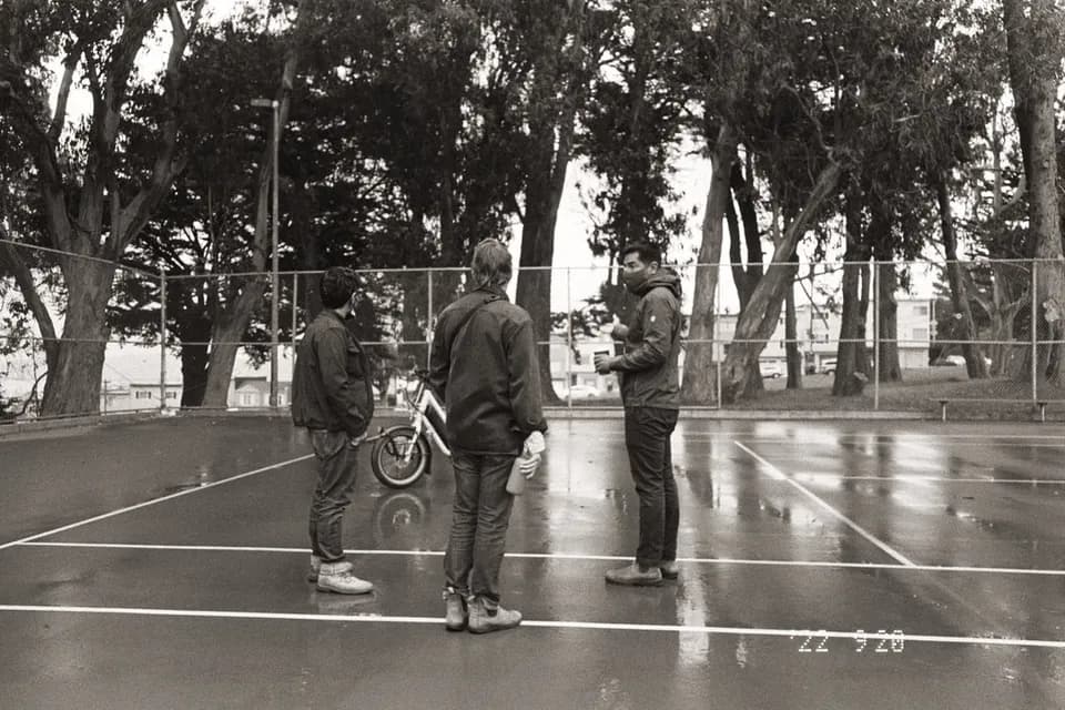 Black and white BTS photo of three people talking on a wet tennis court under trees, with a white e-bike behind them