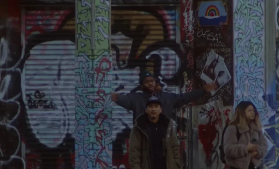 Frame from the Ovrkast. Outro (It's Time) video: a man with arms outstretched before a graffiti wall, two people in front