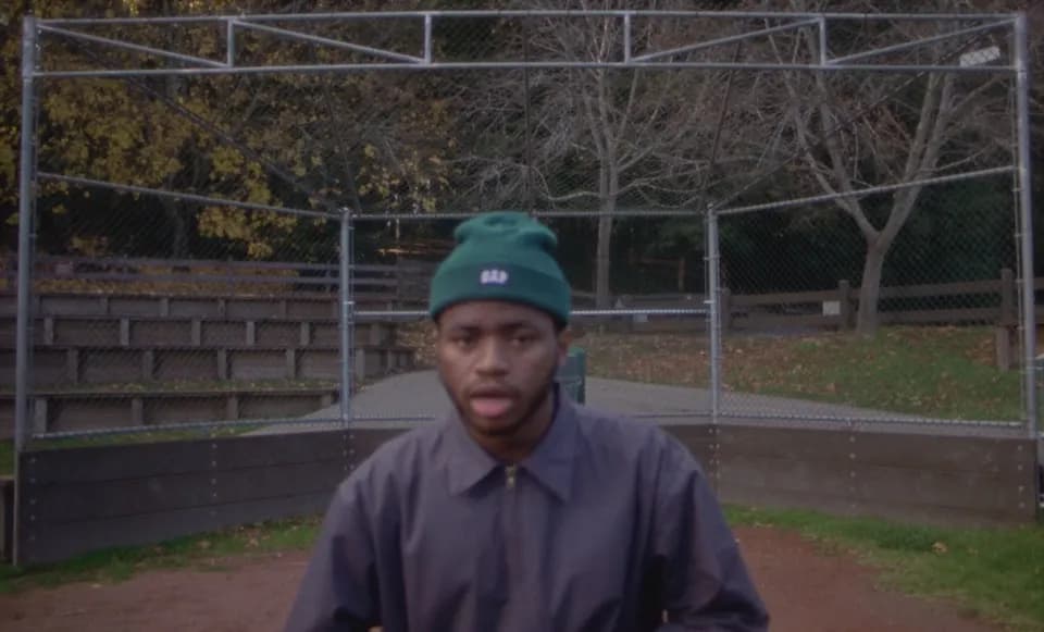 Man in a green beanie and navy zip jacket facing the camera in front of a chain-link backstop at a baseball field