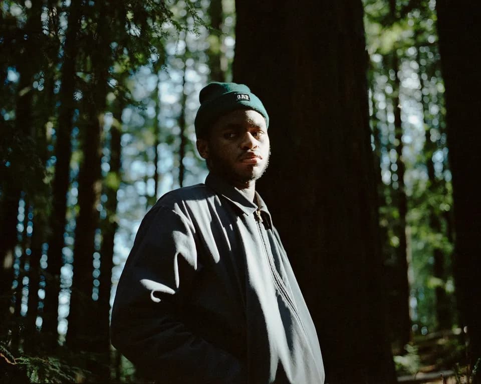 Portrait of a man in a green beanie and dark zip jacket standing among tall redwood trees in dappled light