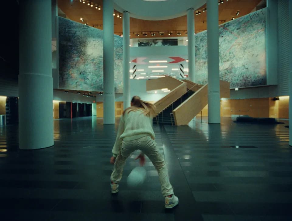 Woman in a cream sweater dribbling a basketball in a museum atrium with large murals, columns, and a wooden staircase