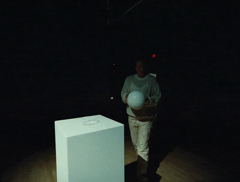 Woman in a cream sweater holding a white basketball beside a white plinth in a dark gallery