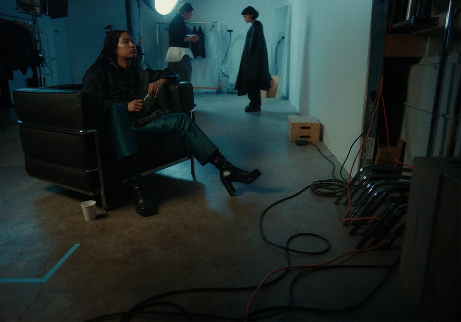 Woman in black boots seated in a leather armchair on a teal-lit set, with crew and a person in a cape behind her