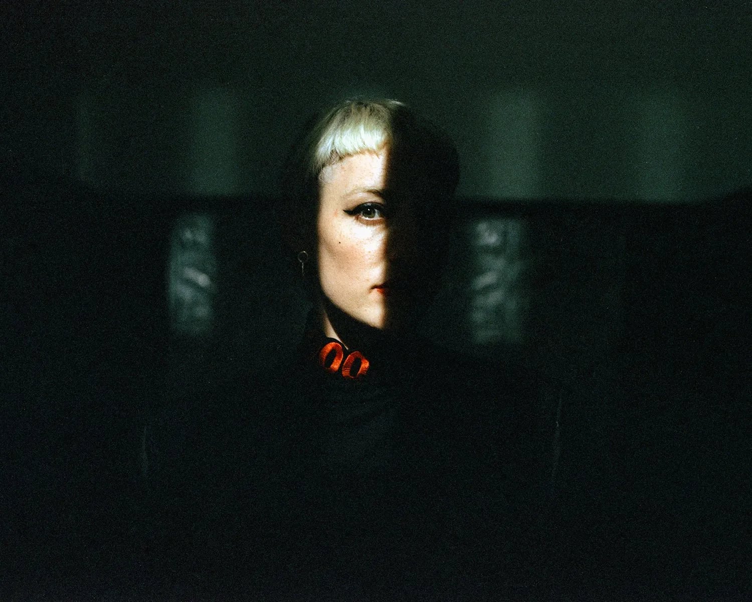 Portrait of a woman with platinum bangs, half her face lit by a sliver of light against a dark green background