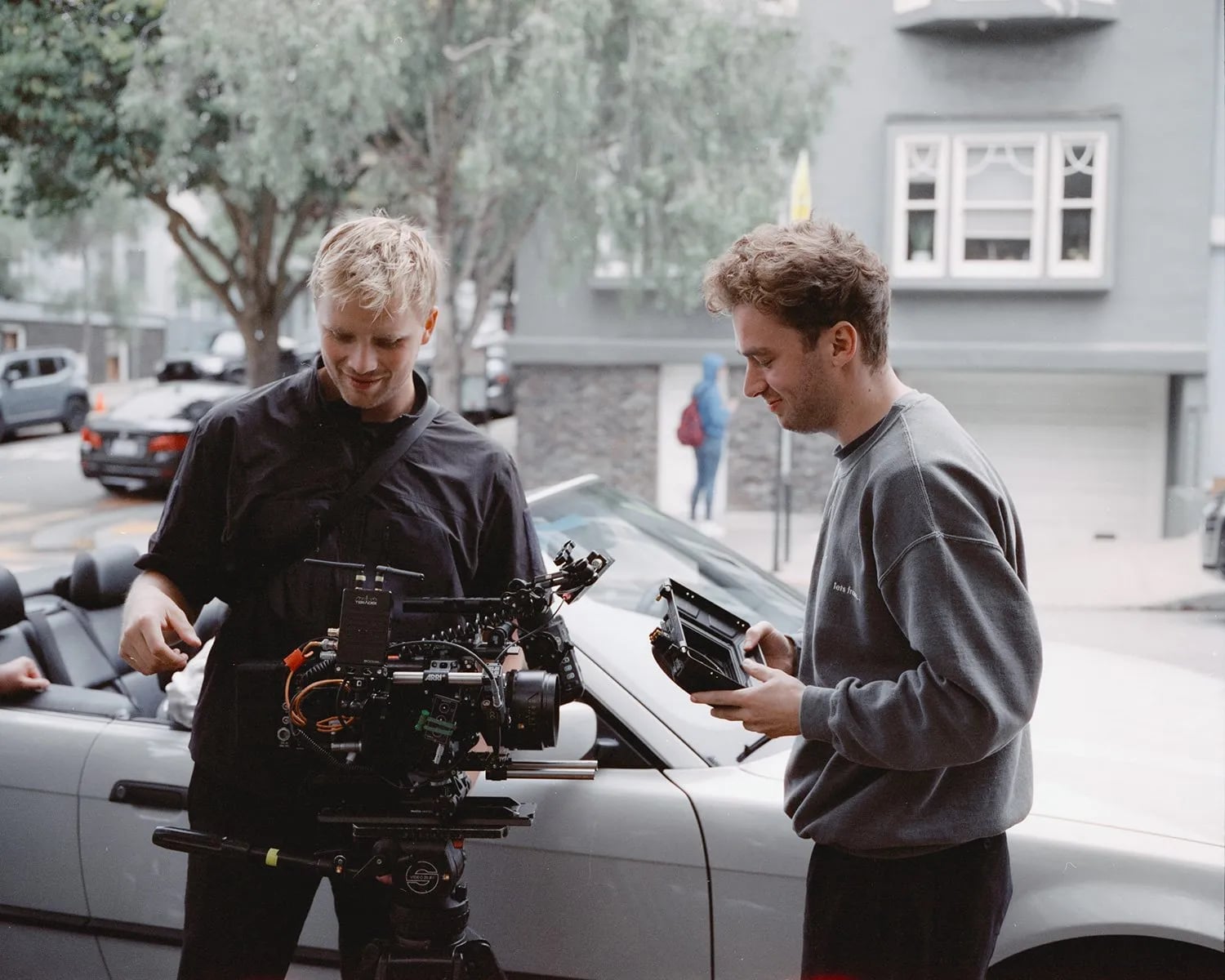 Two men setting up a cinema camera on a tripod beside a silver convertible, one holding a monitor, on a city street
