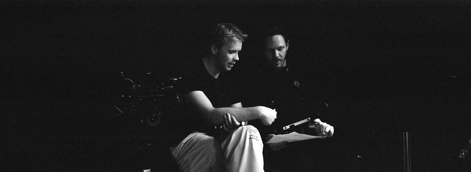 Black and white frame of Skyler and a man in a dark studio looking over a sheet of paper beside camera equipment