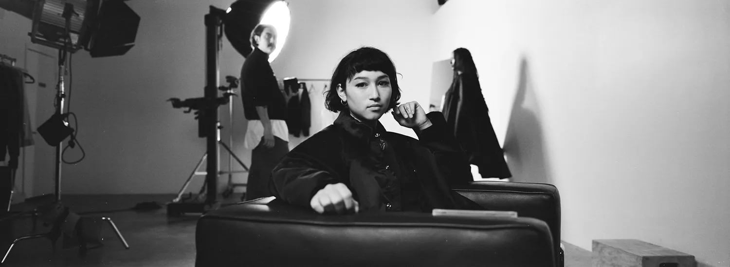 5000 film behind the scenes: woman with a short bob and bangs in a black leather armchair, studio lights behind