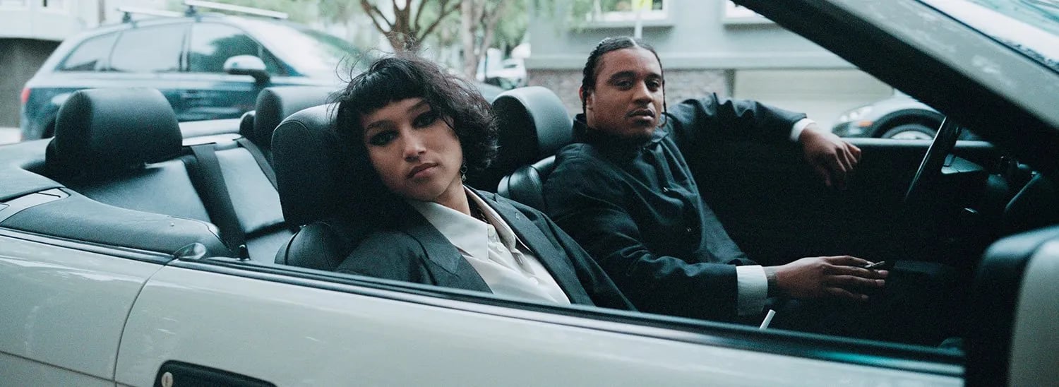 Behind the scenes on film: a woman and a man in dark jackets sitting in a silver convertible on a city street
