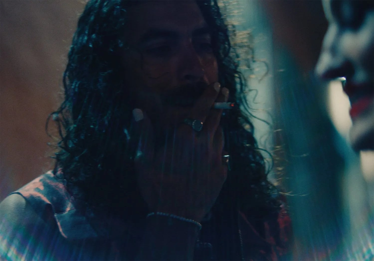 Close, dark shot of a man with long curly hair and round glasses holding a cigarette, a blurred face at right