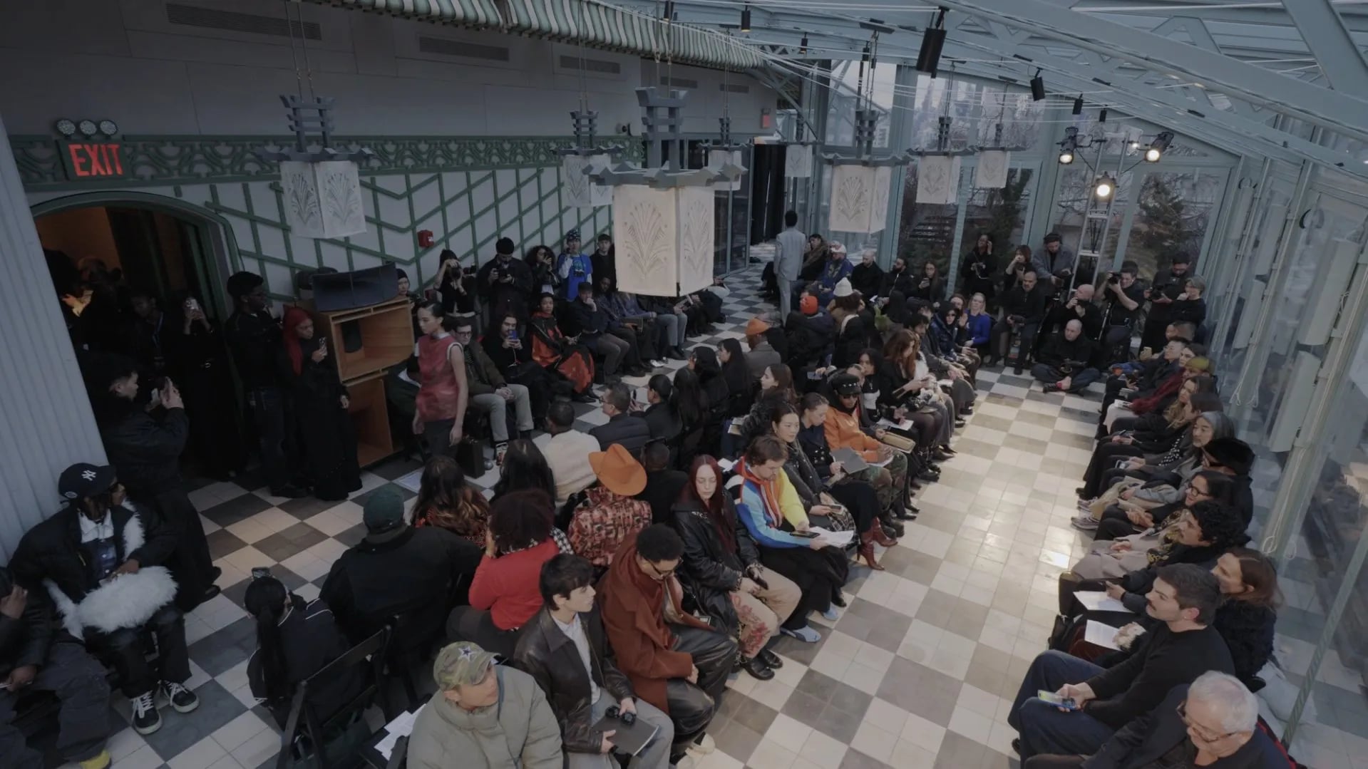 High-angle view of the runway audience on both sides of a checkerboard aisle under an exit sign and paper lanterns