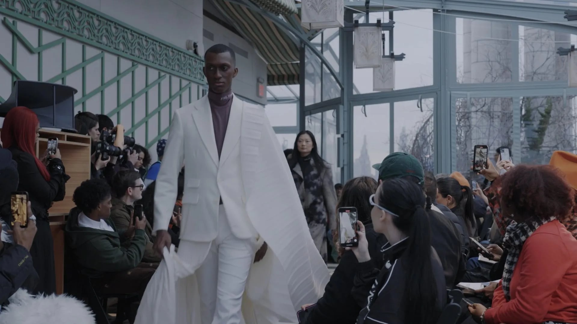 5000 Runway Edit: model in an all-white suit with a pleated white cape, guests holding phones at right