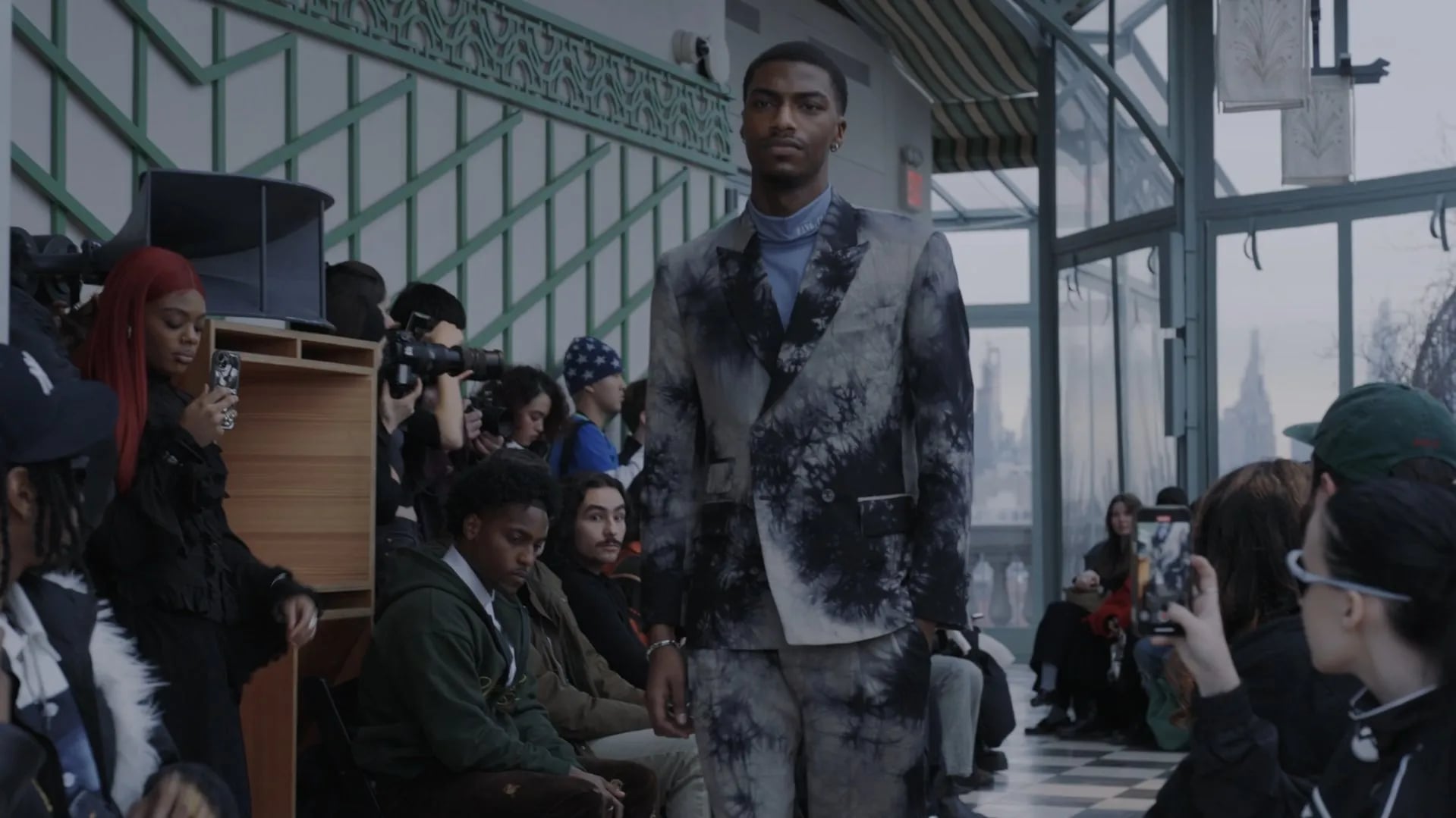 Model in a black and grey tie-dyed suit over a blue turtleneck, with photographers and guests behind