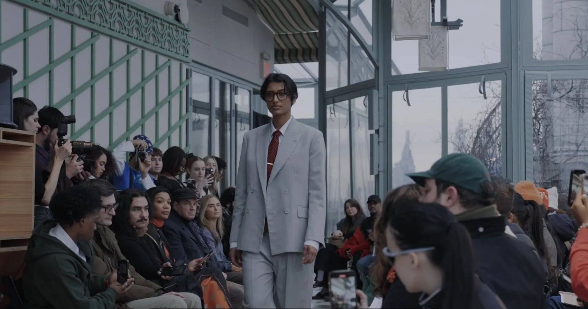 5000 Runway Edit: model in a grey double-breasted suit and red tie walks past photographers and seated guests