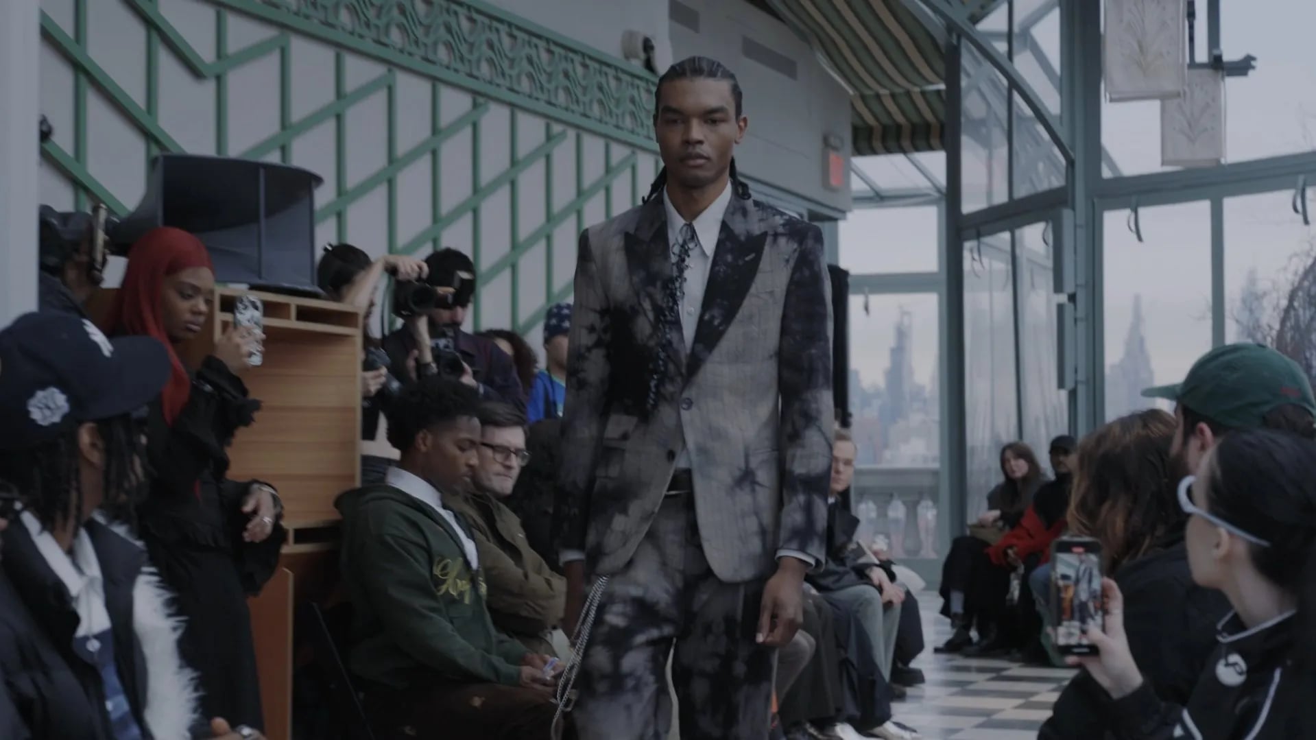 Runway at the 5000 Bootsy Collection show: model in a grey tie-dyed suit and patterned tie, red-haired guest at left