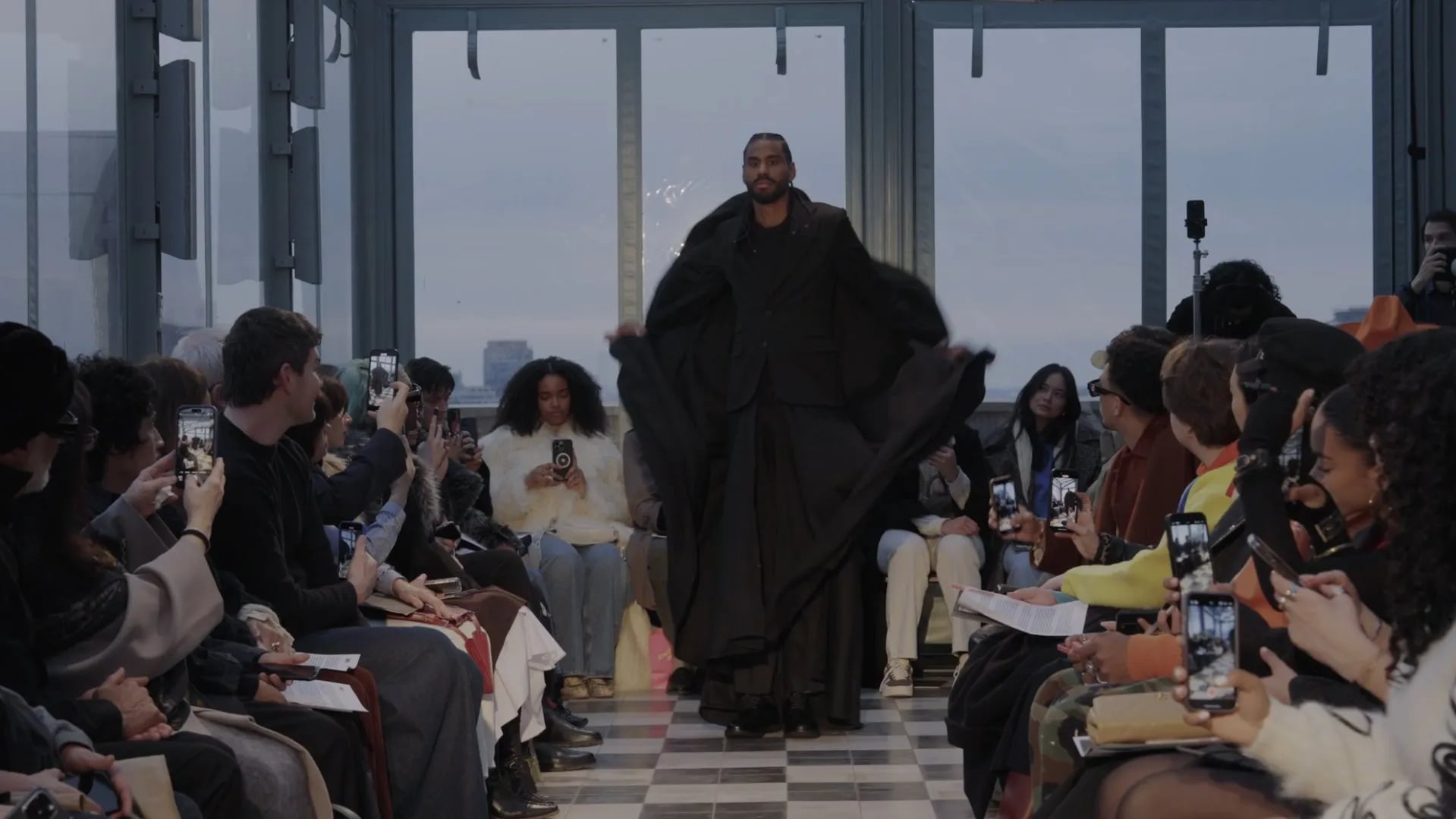 Model in a long black cape-like coat with arms spread, walking toward camera as guests film on phones