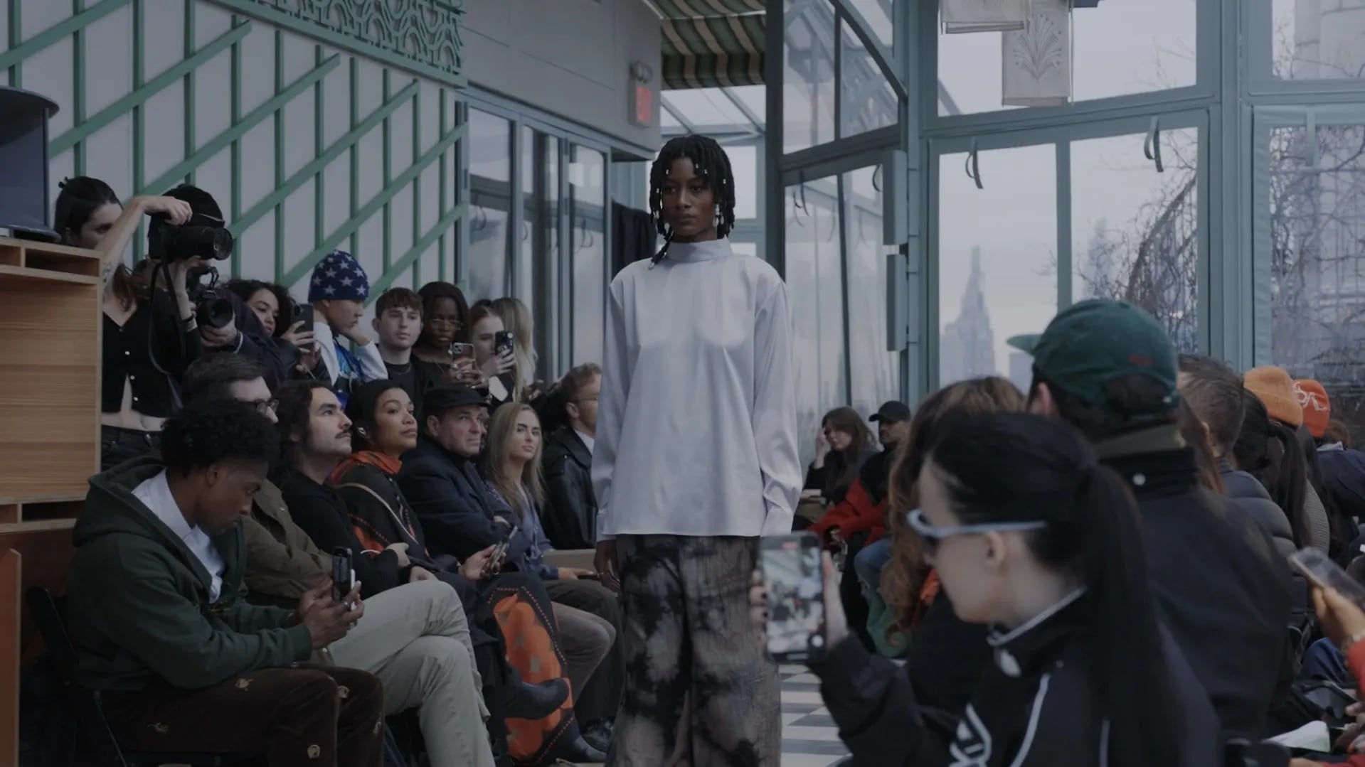 Model in a pale grey high-neck top and tie-dyed trousers, framed between seated guests and a phone in the foreground