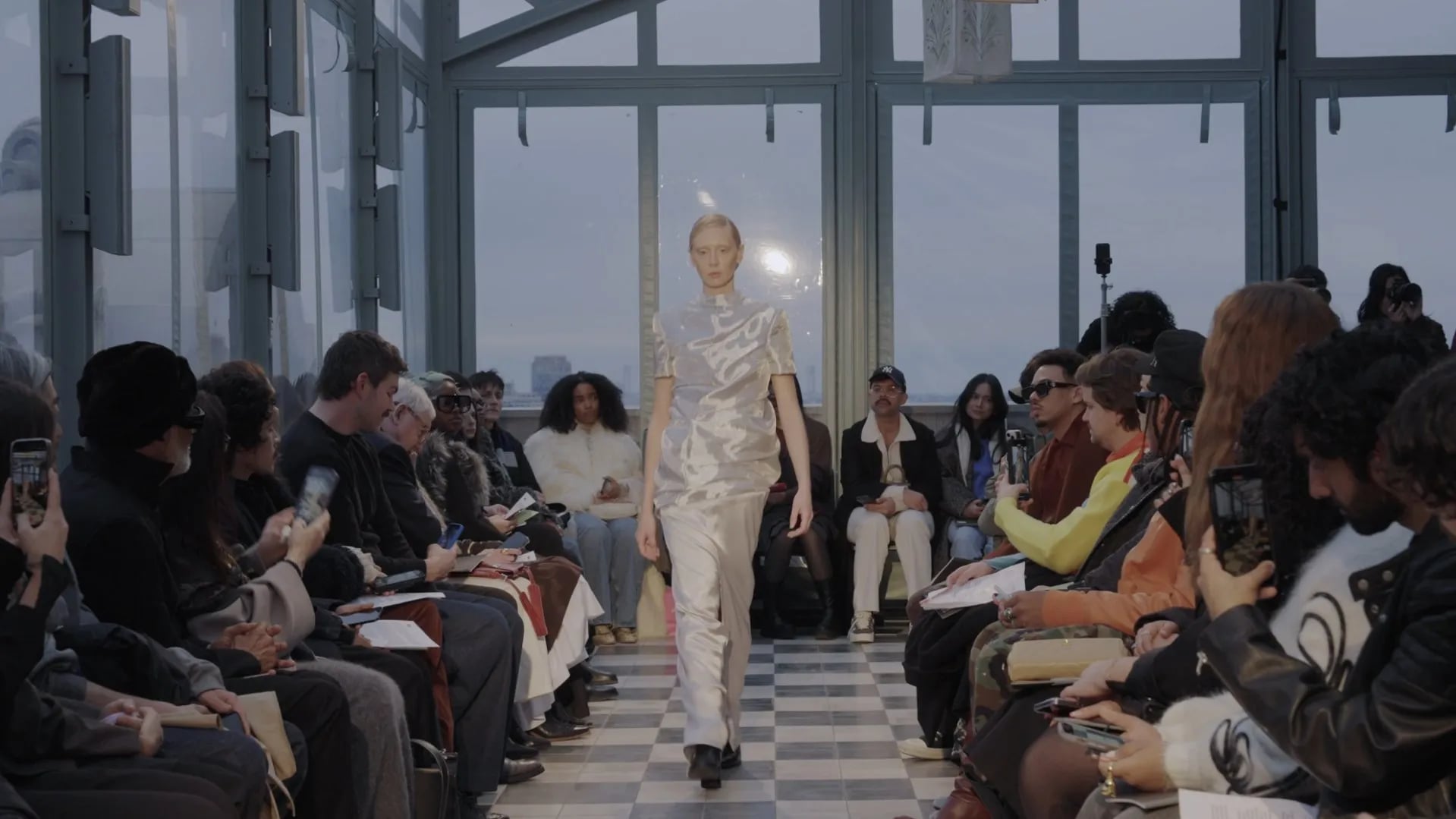 Model in a shimmering silver top and trousers walking the checkerboard runway, with guests filming on both sides
