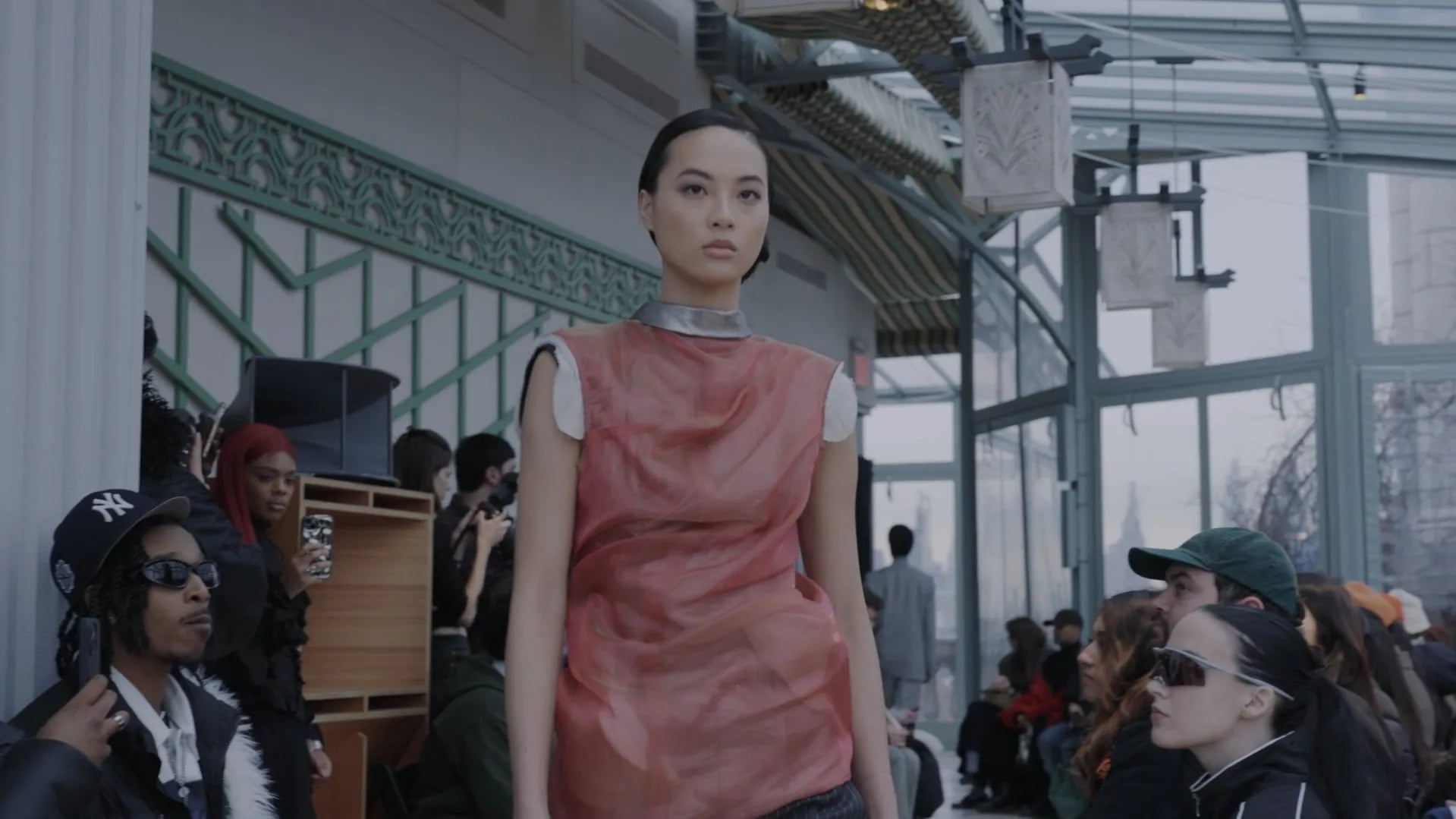 Close shot of a model in a sleeveless red coral sheer top with a silver collar, guests in sunglasses at the edges