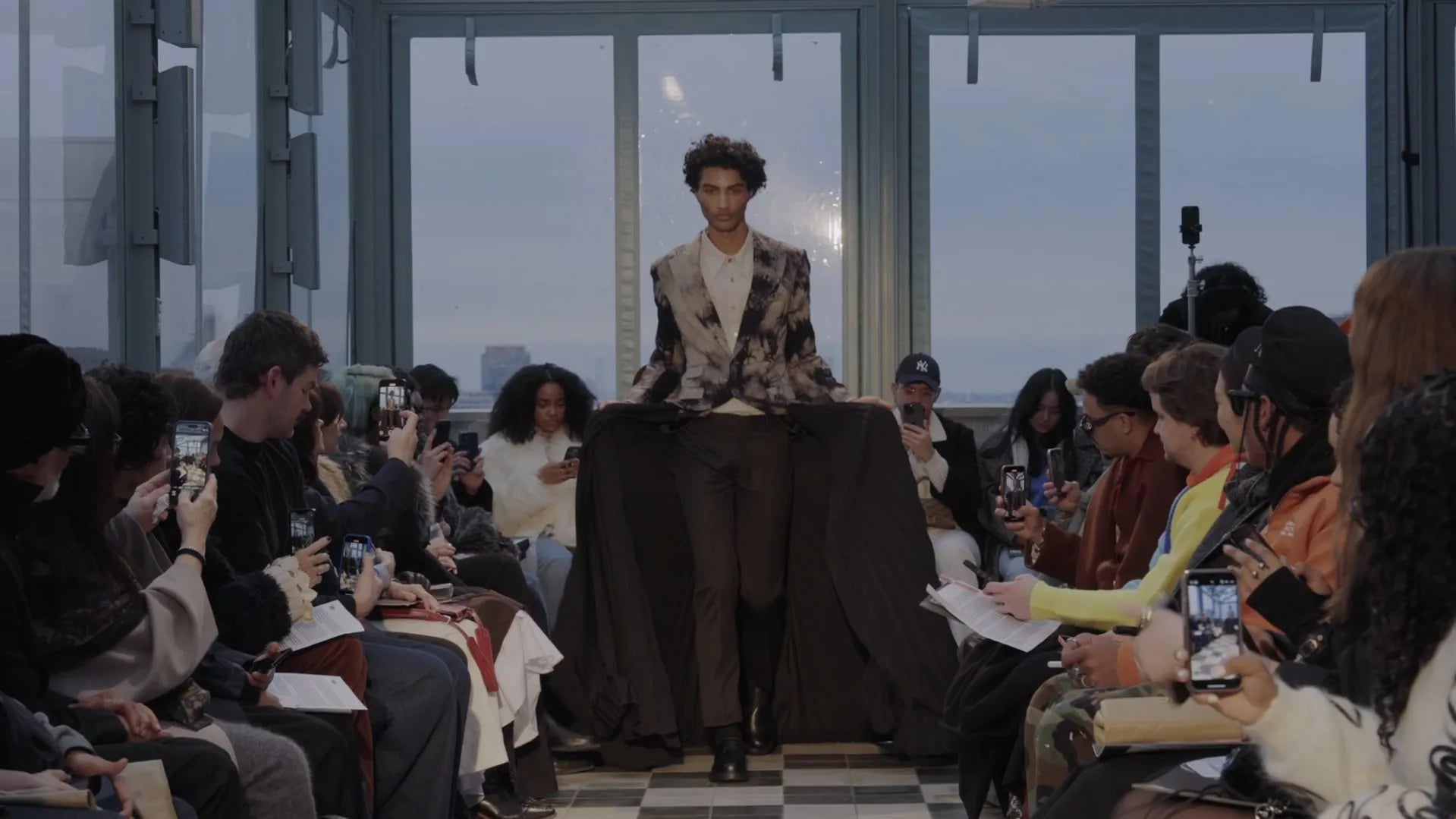 Model in a tie-dyed jacket and brown trousers with a wide dark pleated cape spread behind, guests filming