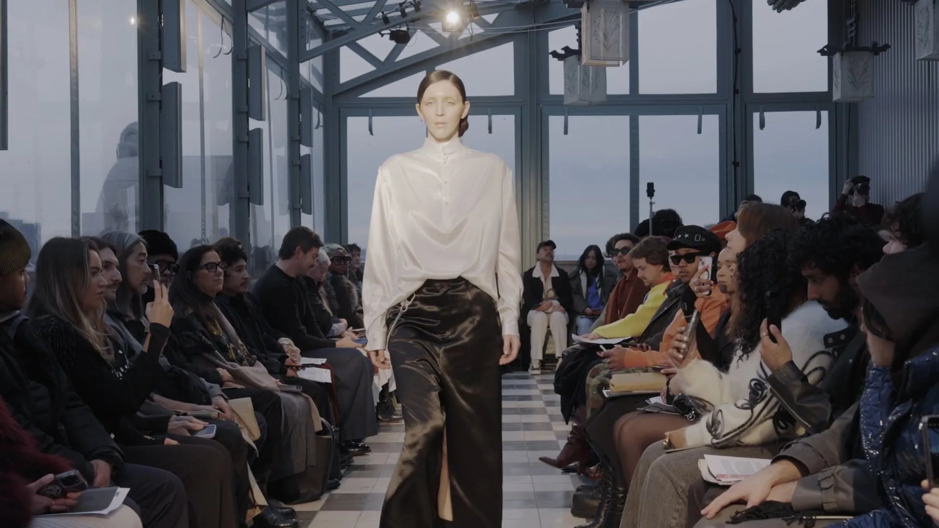Model in a white satin blouse and dark satin trousers walking a checkerboard-floor runway beside a glass wall