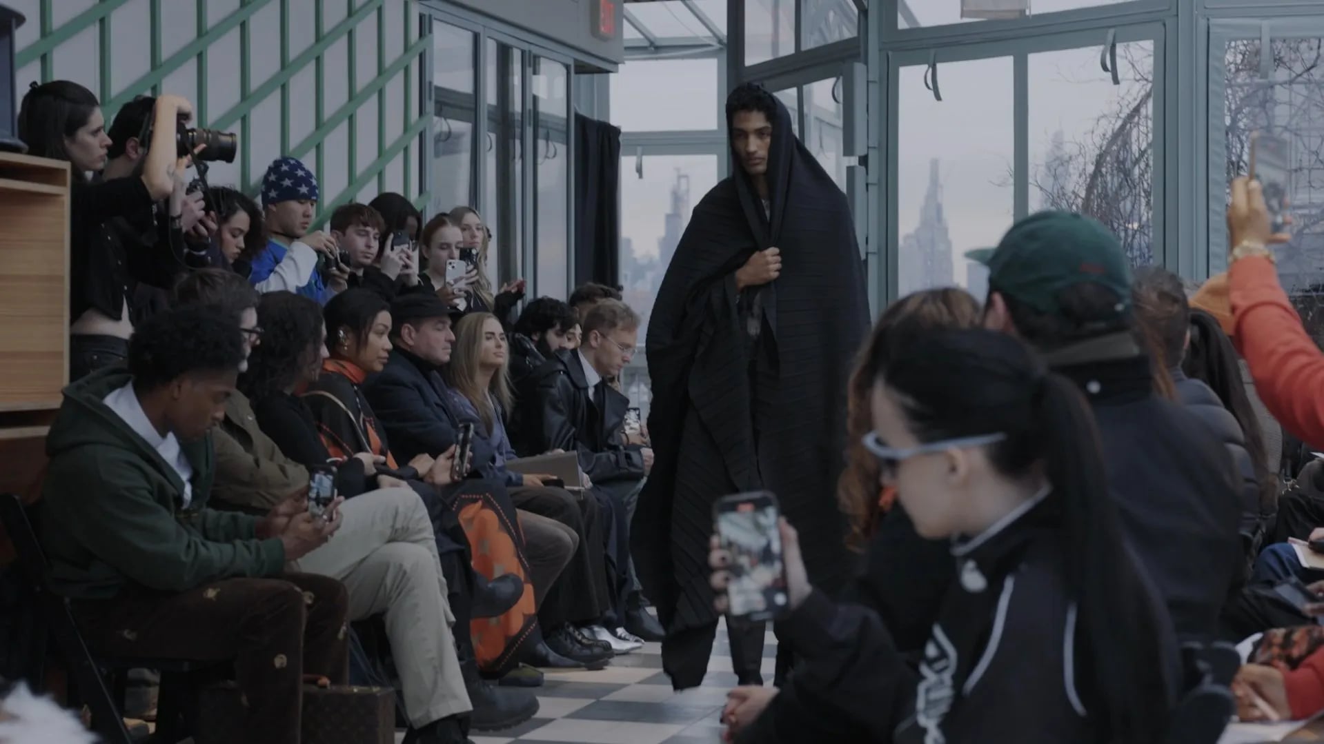 Model wrapped in a black pleated cloak, holding it closed at the chest, seen between rows of seated guests