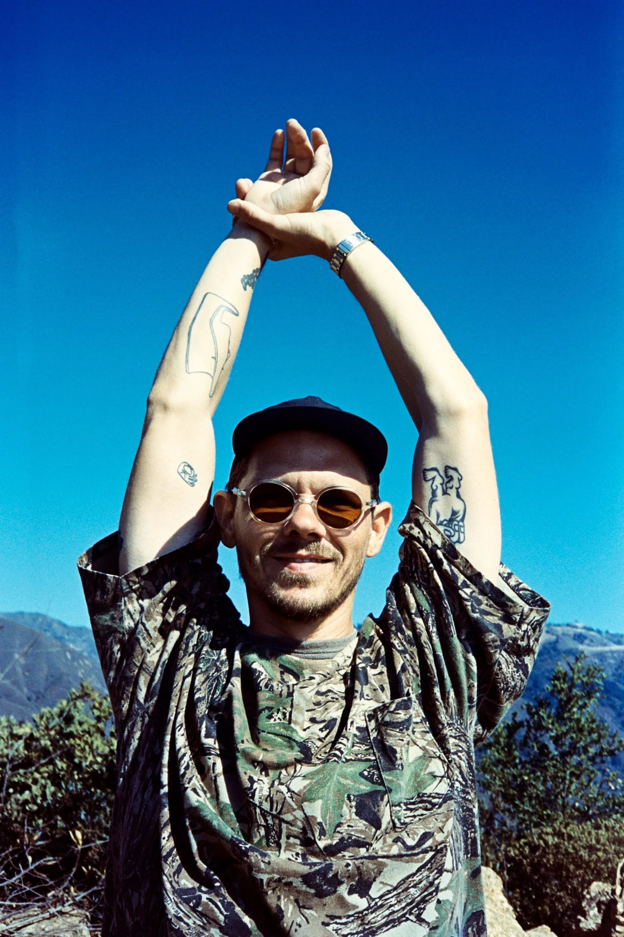 Skyler in sunglasses and a cap with arms raised overhead, wearing a camouflage shirt, against a deep blue sky