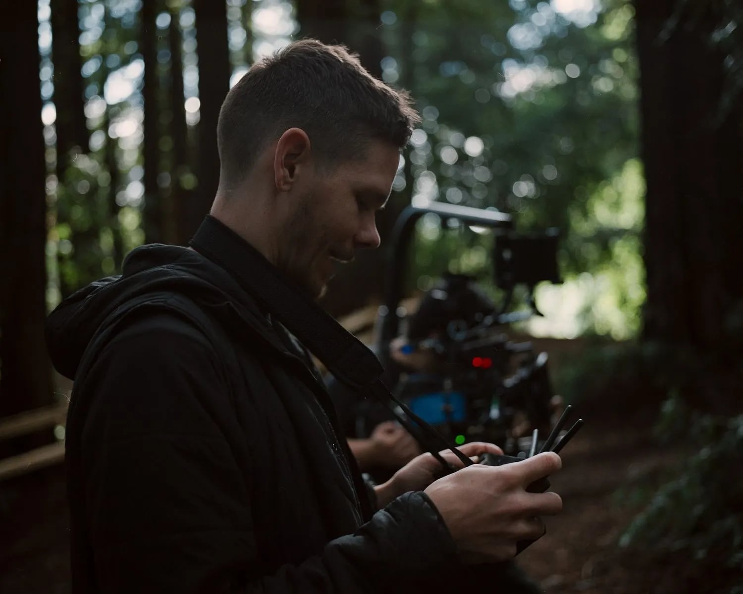 Skyler in a black jacket looks down at a handheld director's monitor in a redwood-like forest, camera rig blurred behind him