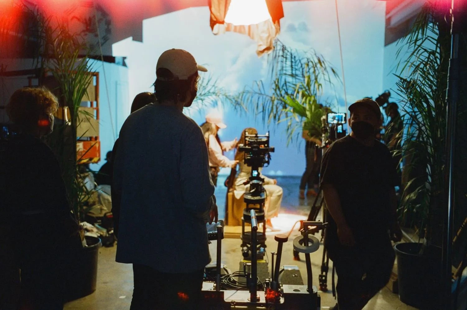 Skyler with film crew and camera on a dolly in a plant-filled studio, with a seated person lit against a cloud backdrop