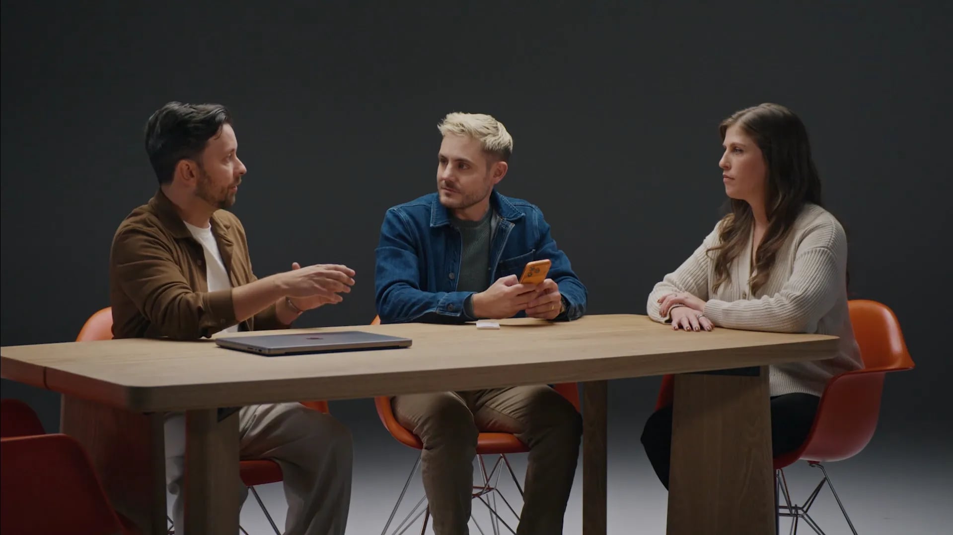 Three people at a wooden table in a grey studio: a man gesturing, a man in denim holding an orange phone, a woman listening