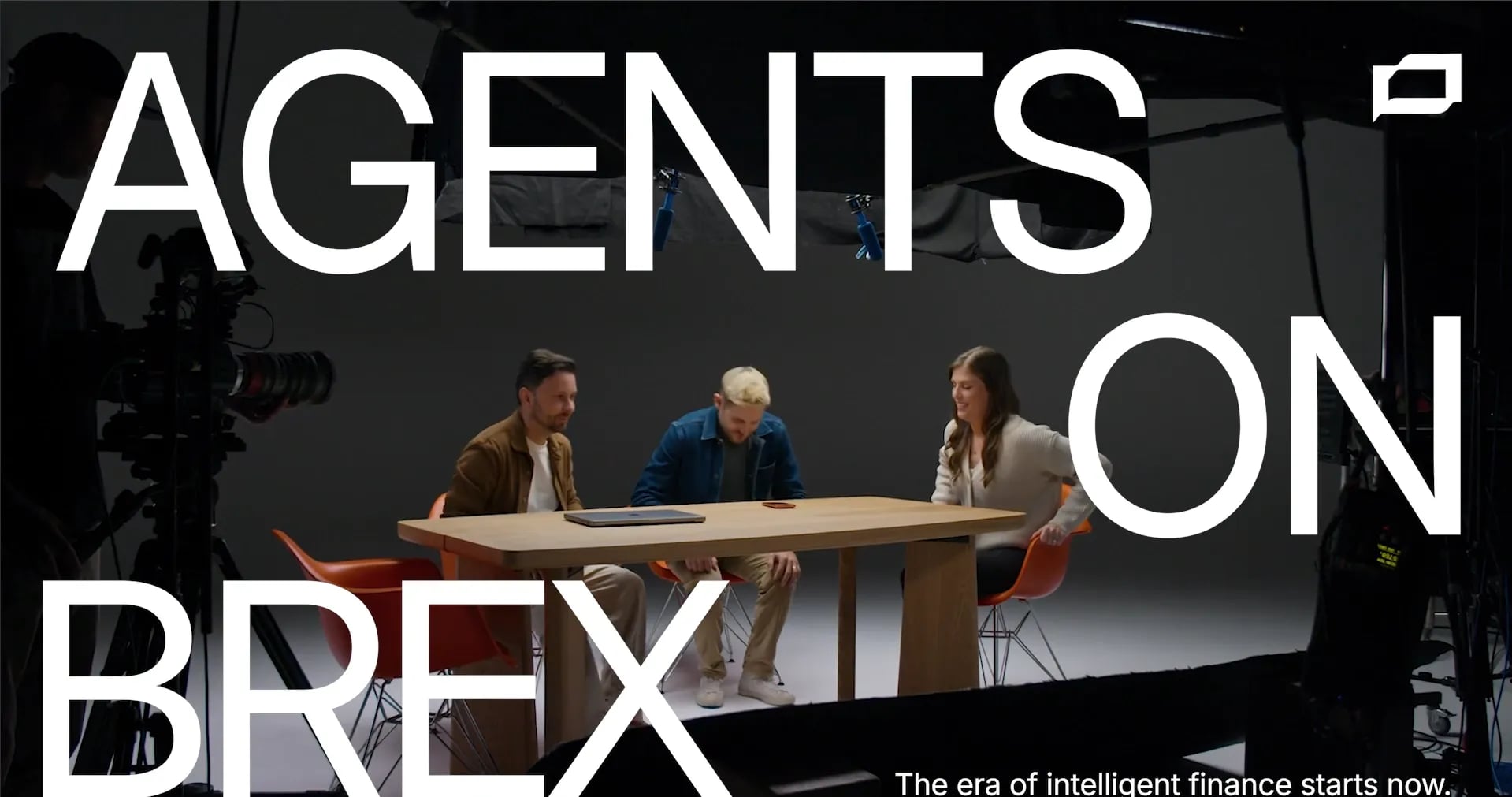Three people at a wooden table on a dark film set, with the white title AGENTS ON BREX across the frame