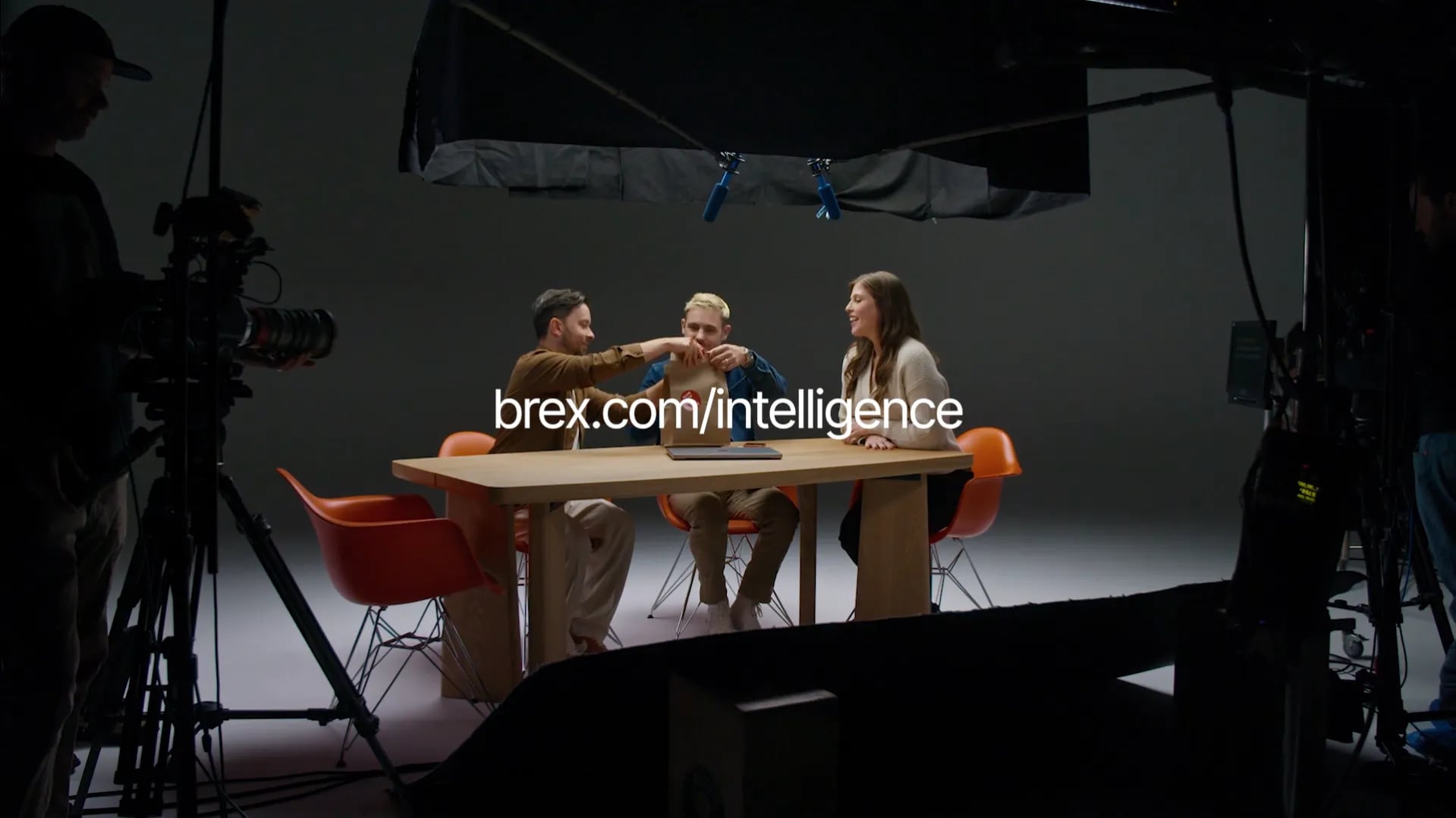 Wide studio shot of three people at a wooden table handling a paper bag, with the text brex.com/intelligence overlaid