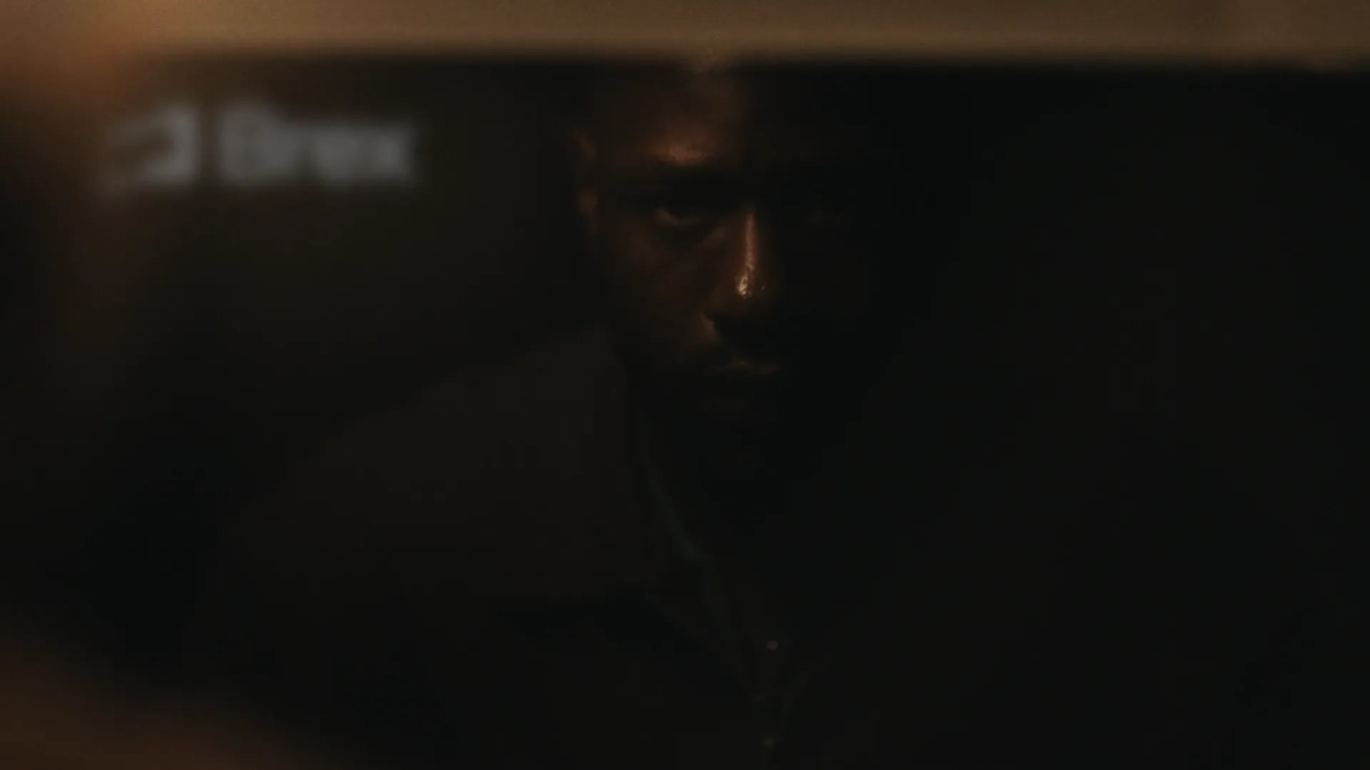 Dim close-up of a man's face half in shadow reflected in a screen with a blurred Brex logo