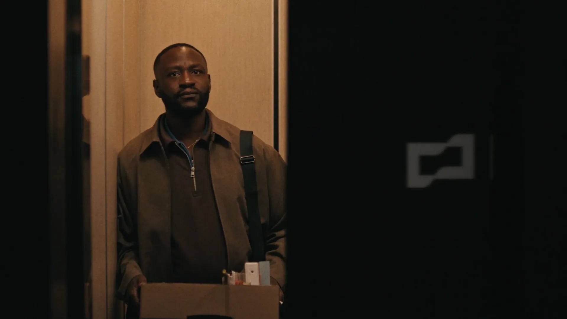 Man in a brown coat holding a cardboard box of belongings in a closing elevator, a Brex logo on the dark wall beside him