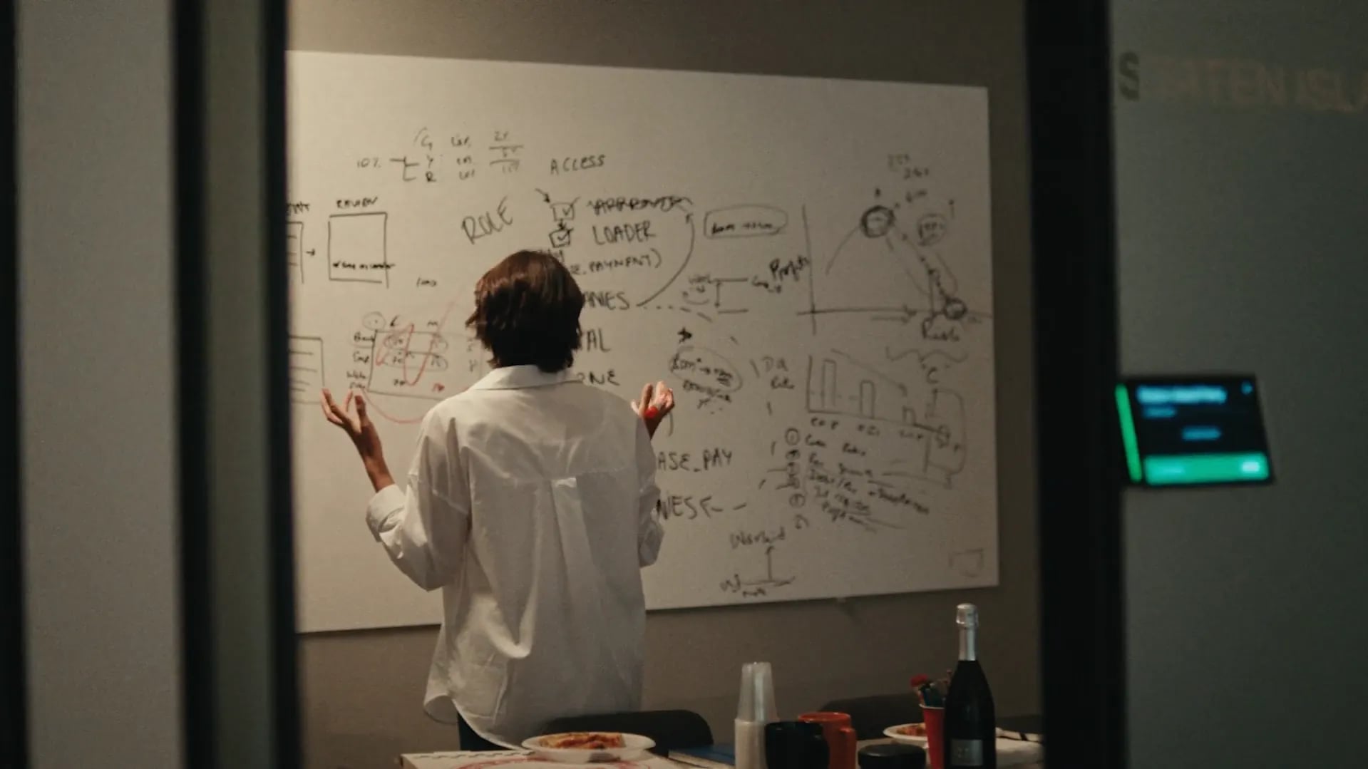 A person in a white shirt with arms raised at a whiteboard covered in diagrams, a pizza and champagne bottle on the table