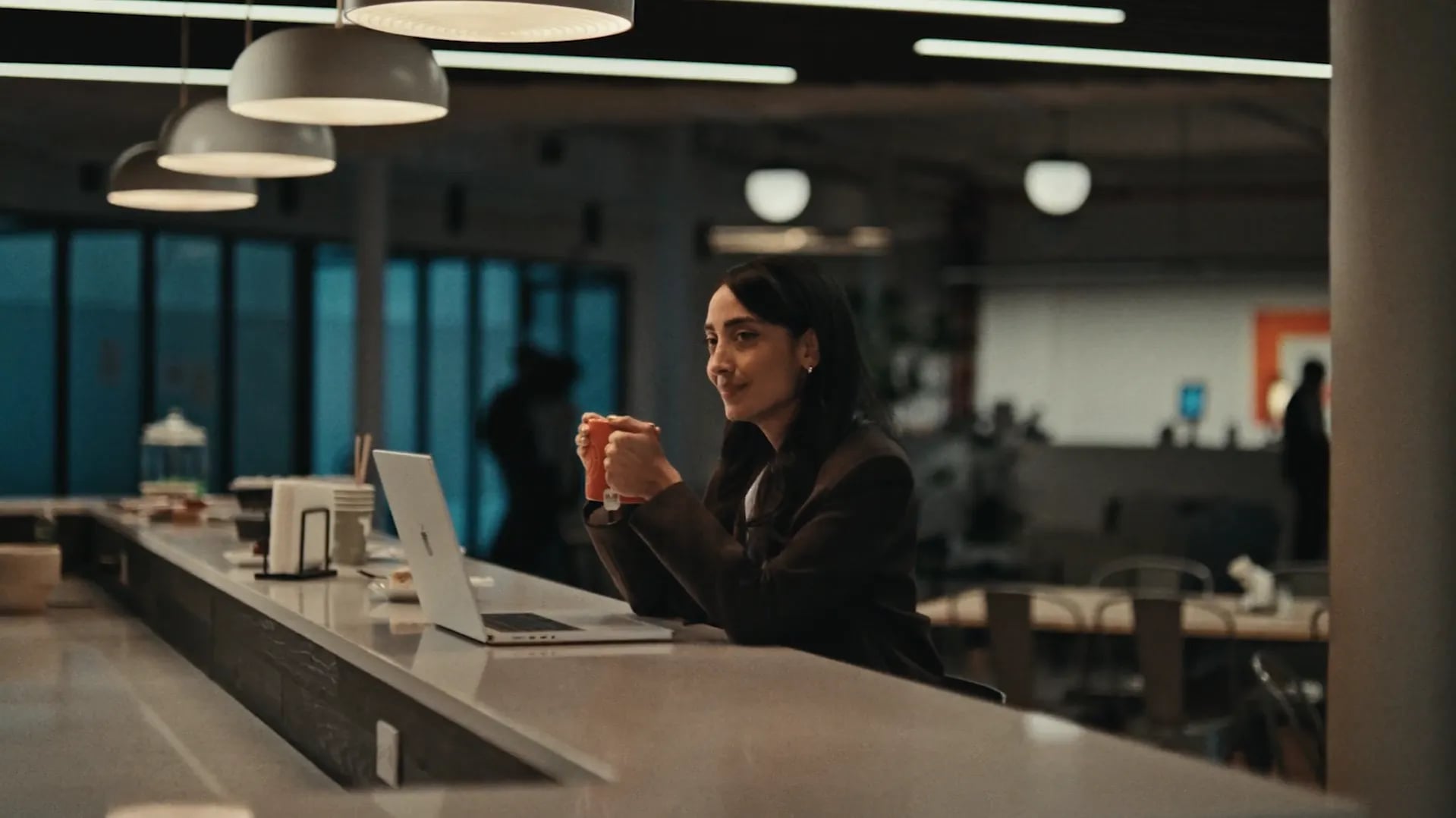 Woman in a dark blazer holding an orange mug at a long counter beside a laptop in a dim open-plan office