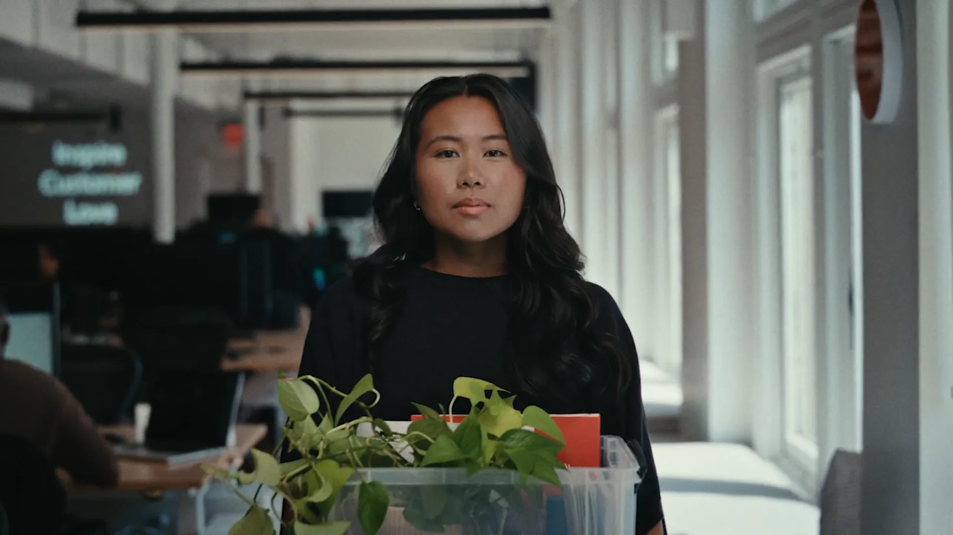 Woman in a black top carrying a clear box with a green plant and red folder down a bright office hallway