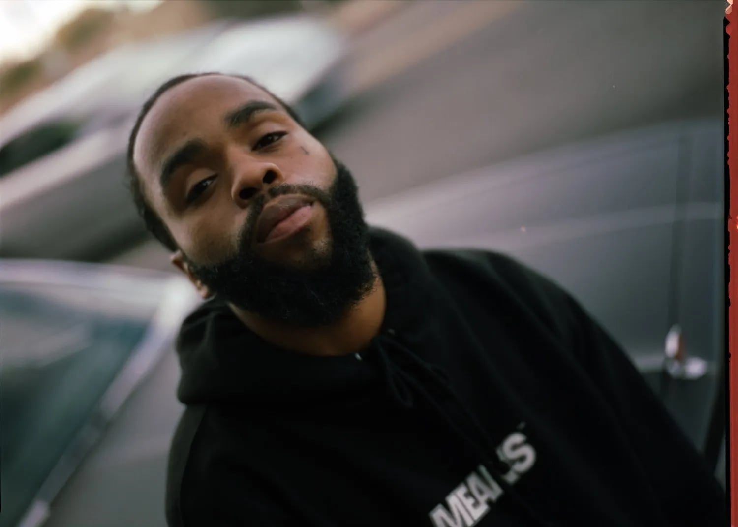Fly Anakin No Dough music video still: bearded man in a black hoodie leaning toward the camera beside a parked car